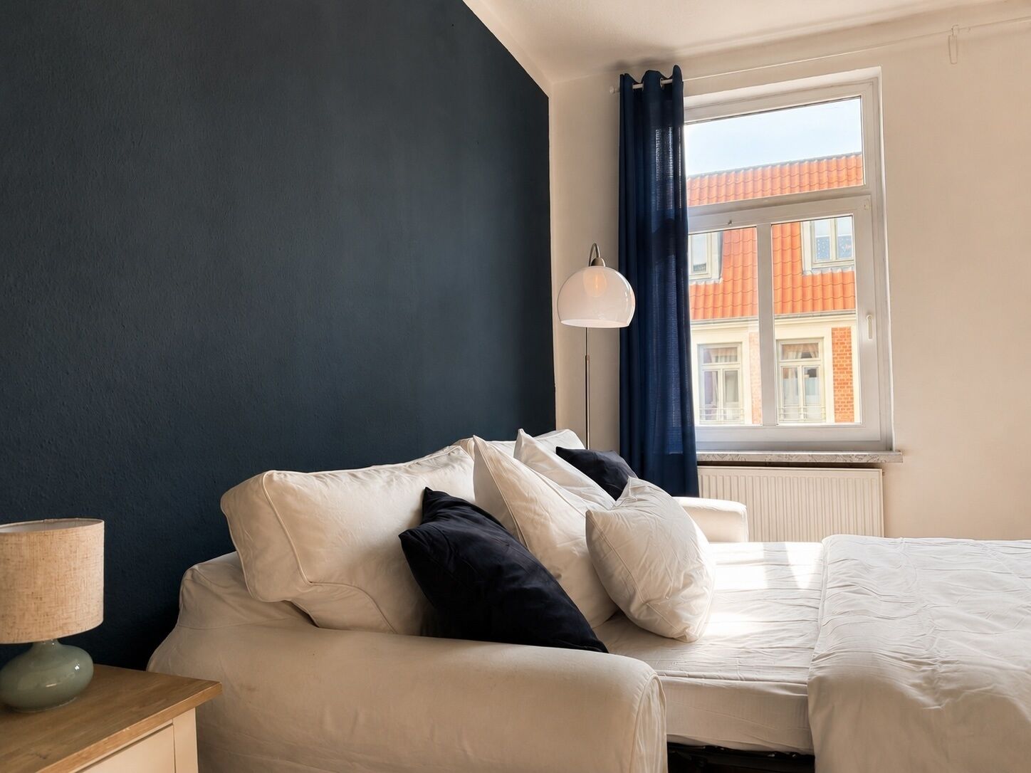A bedroom with a bed, a bedside table with a lamp, a window with curtains, and a view of buildings outside.