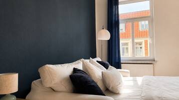 A bedroom with a bed, a bedside table with a lamp, a window with curtains, and a view of buildings outside.