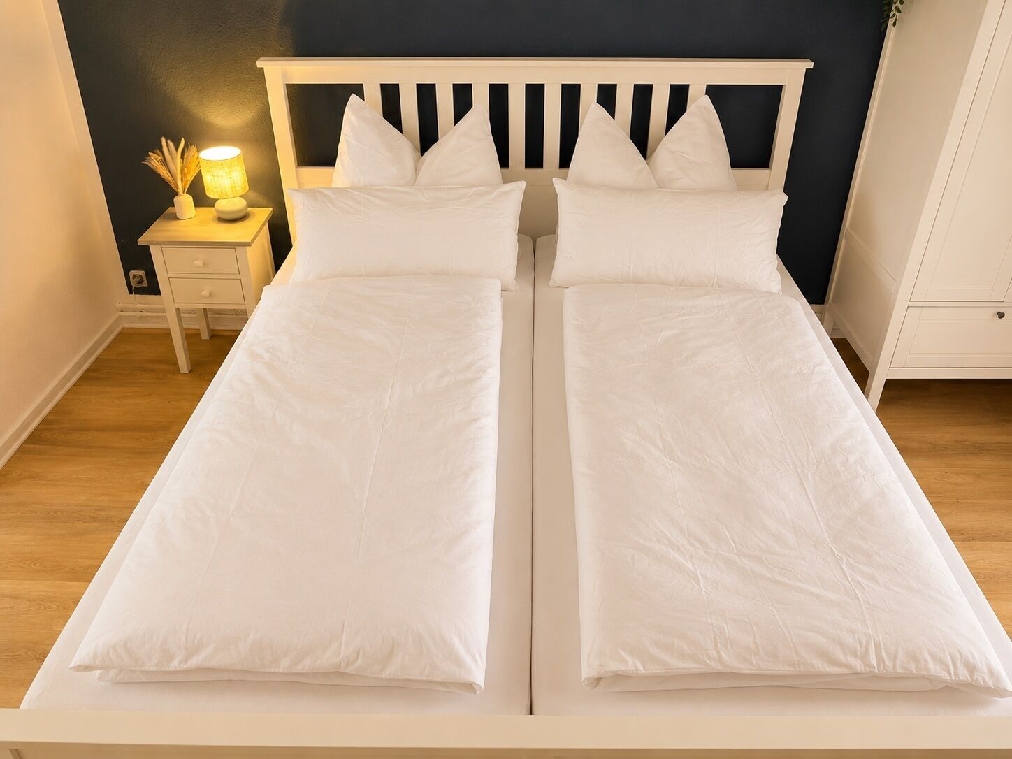 Ein Doppelbett mit weißer Bettwäsche, ein Nachttisch mit Lampe und eine dunkle Wand.