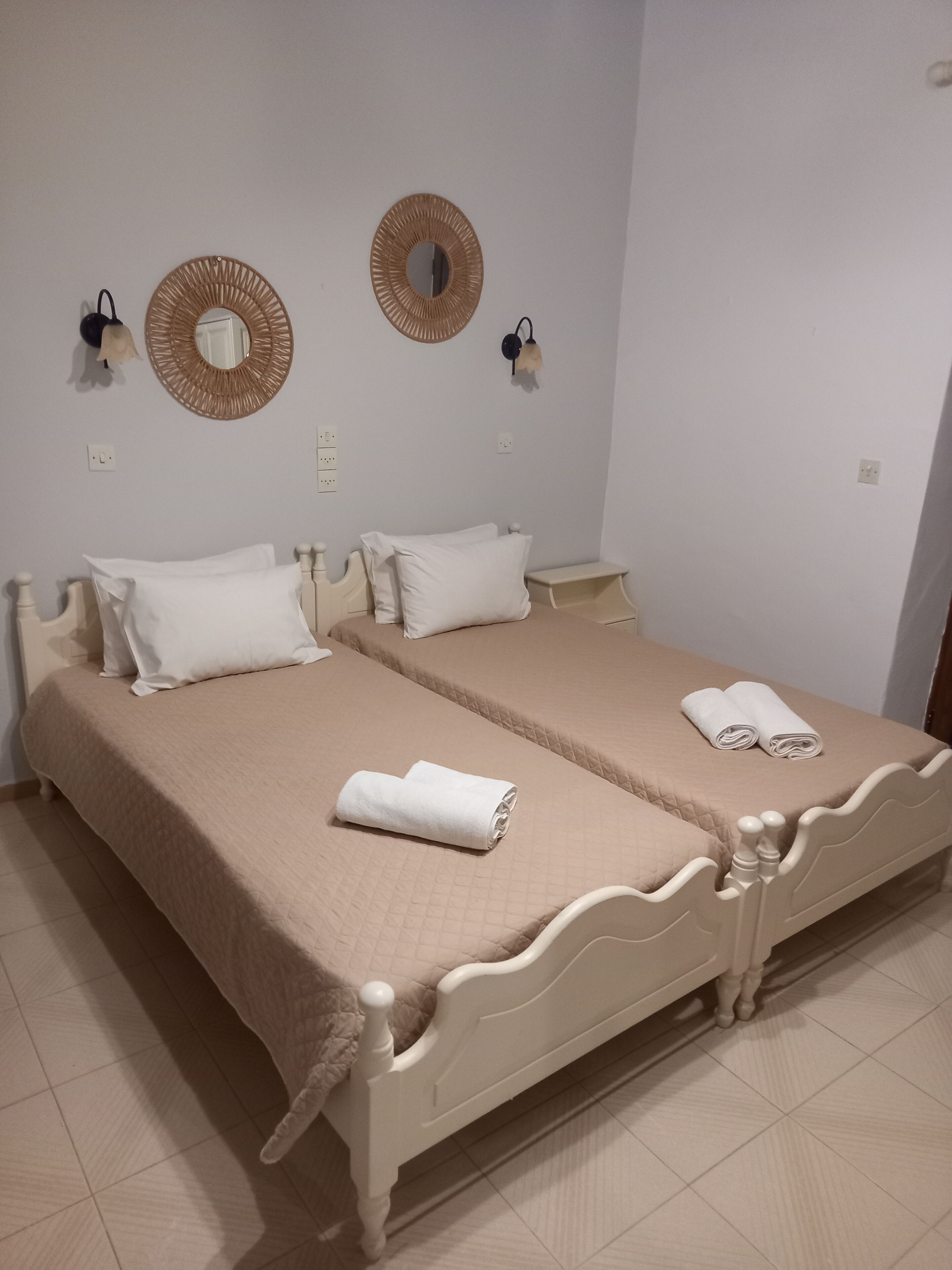 A double bed with a beige bedspread, two pillows, and two rolled towels. There are two wall-mounted sconces with decorative mirrors above the bed.