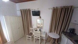 A room with a desk, chair, TV, mirror, and kitchenette. - DIAMOND VIEW (Lesvos)