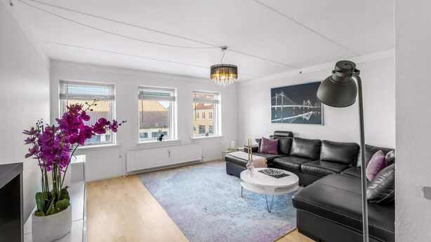 Ett modernt vardagsrum med en svart lädersoffa, ett runt soffbord, en golvlampa och ett stort fönster med persienner.