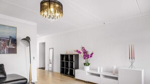 Ett modernt vardagsrum med en vit soffa, ett runt soffbord, en kristallkrona, en tavla som föreställer en stadsbild, ett svart skåp och ett vitt konsolbord med en vas med lila blommor.