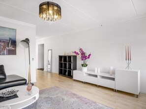 A modern living room with a white sofa, a round coffee table, a chandelier, a cityscape painting, a black cabinet, and a white console table with a vase of purple flowers. - 4 Star Holiday Home in Lokken (Lokken)