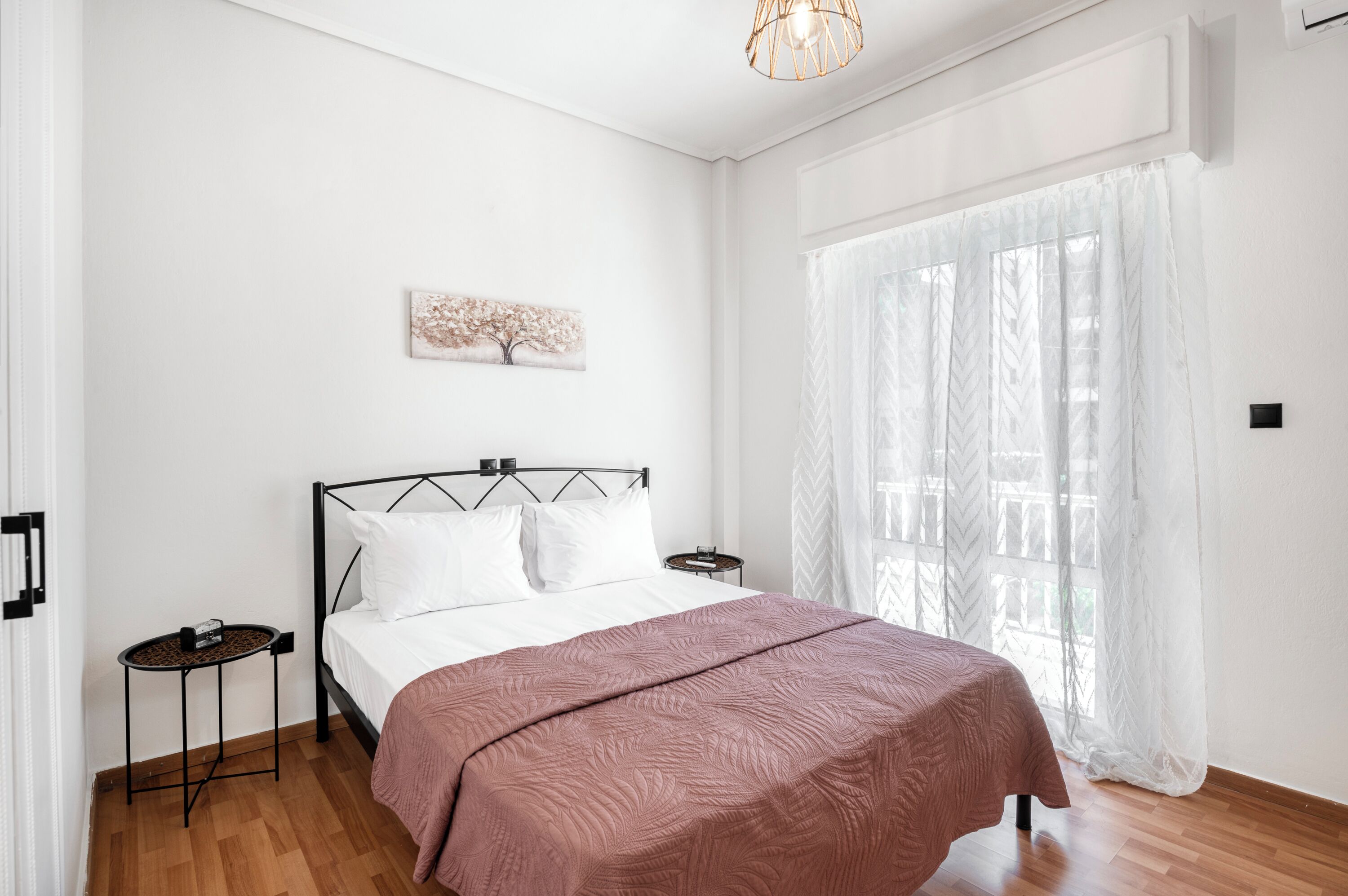 Una camera da letto con un letto, un comodino, un'opera d'arte appesa al muro e una finestra con tende leggere.