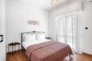 A bedroom with a bed, a bedside table, a wall-mounted artwork, and a window with sheer curtains. - Glyfada Stay (Glifada)
