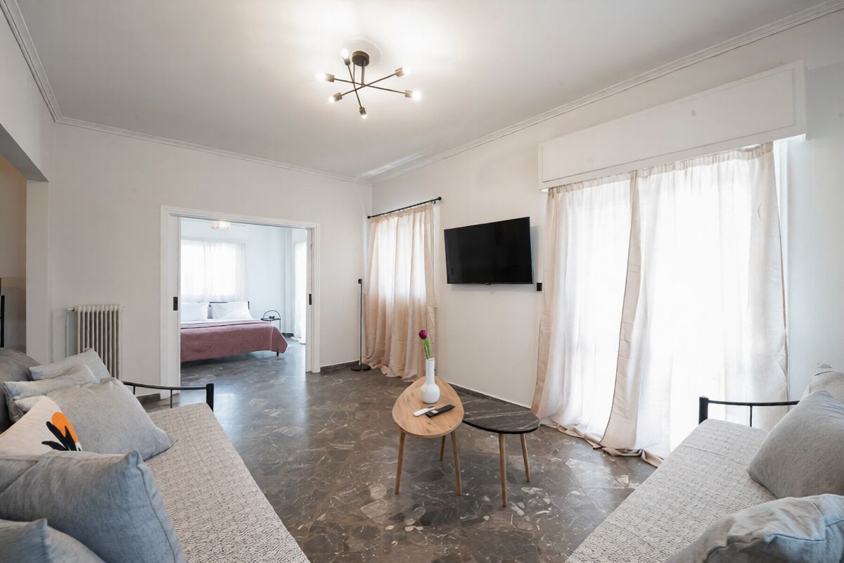A modern living room with a sofa, a coffee table, a TV mounted on the wall, and a view into the adjacent bedroom.