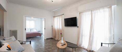 A modern living room with a sofa, a coffee table, a TV mounted on the wall, and a view into the adjacent bedroom.