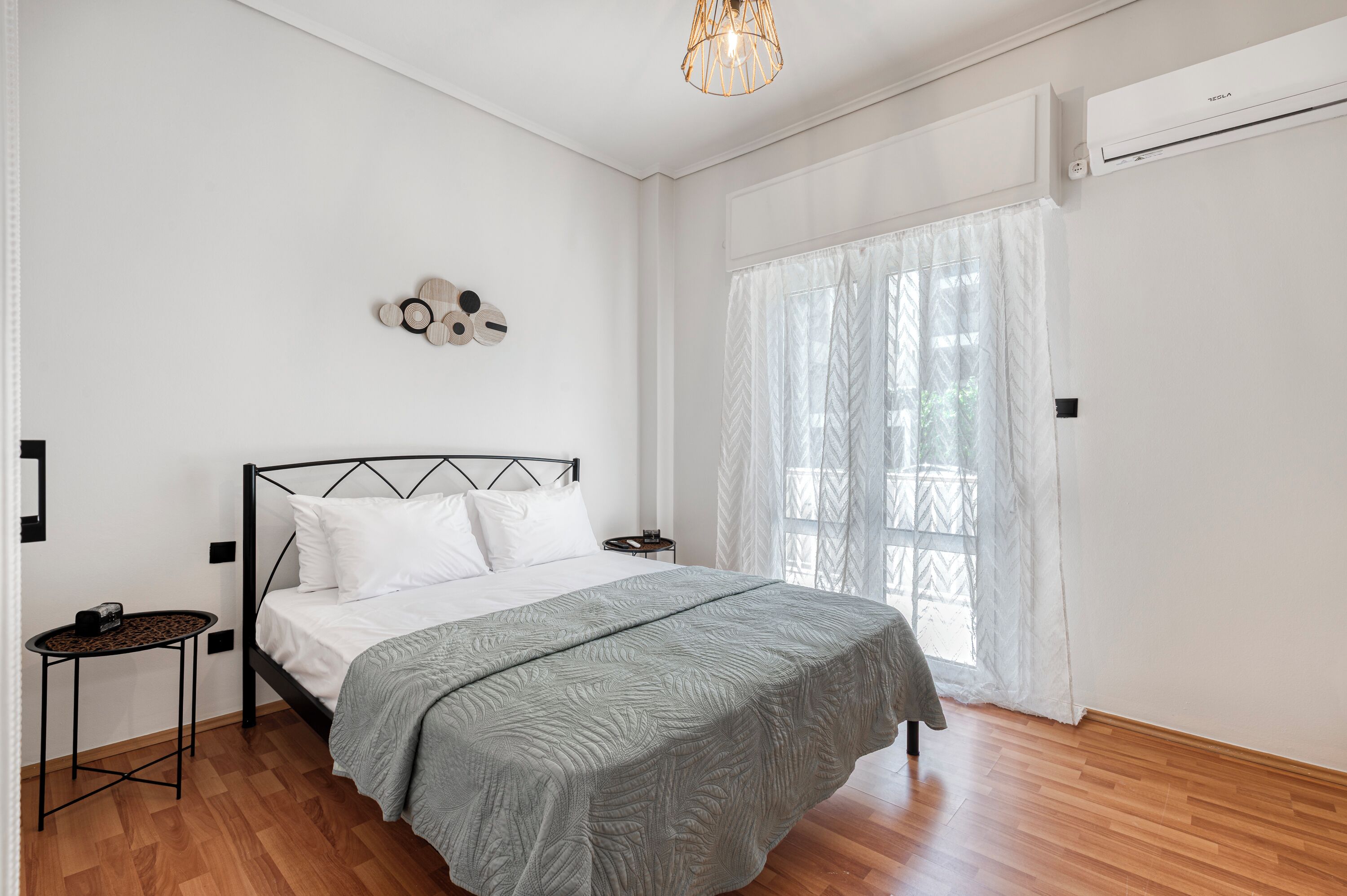 A bedroom with a bed, a bedside table, a wall-mounted light fixture, and a window with sheer curtains.