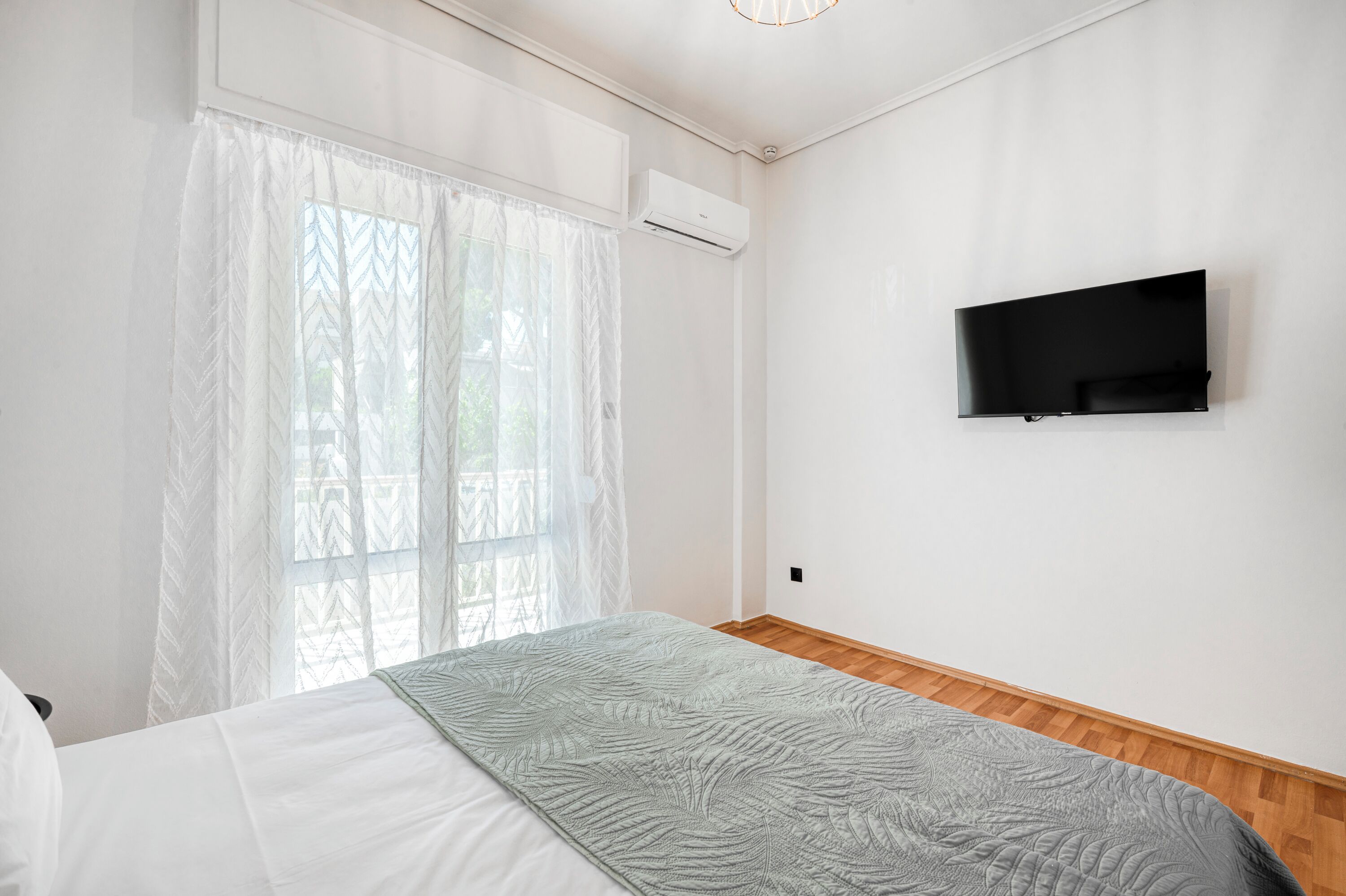 A bedroom with a bed, a TV mounted on the wall, and a window with sheer curtains.