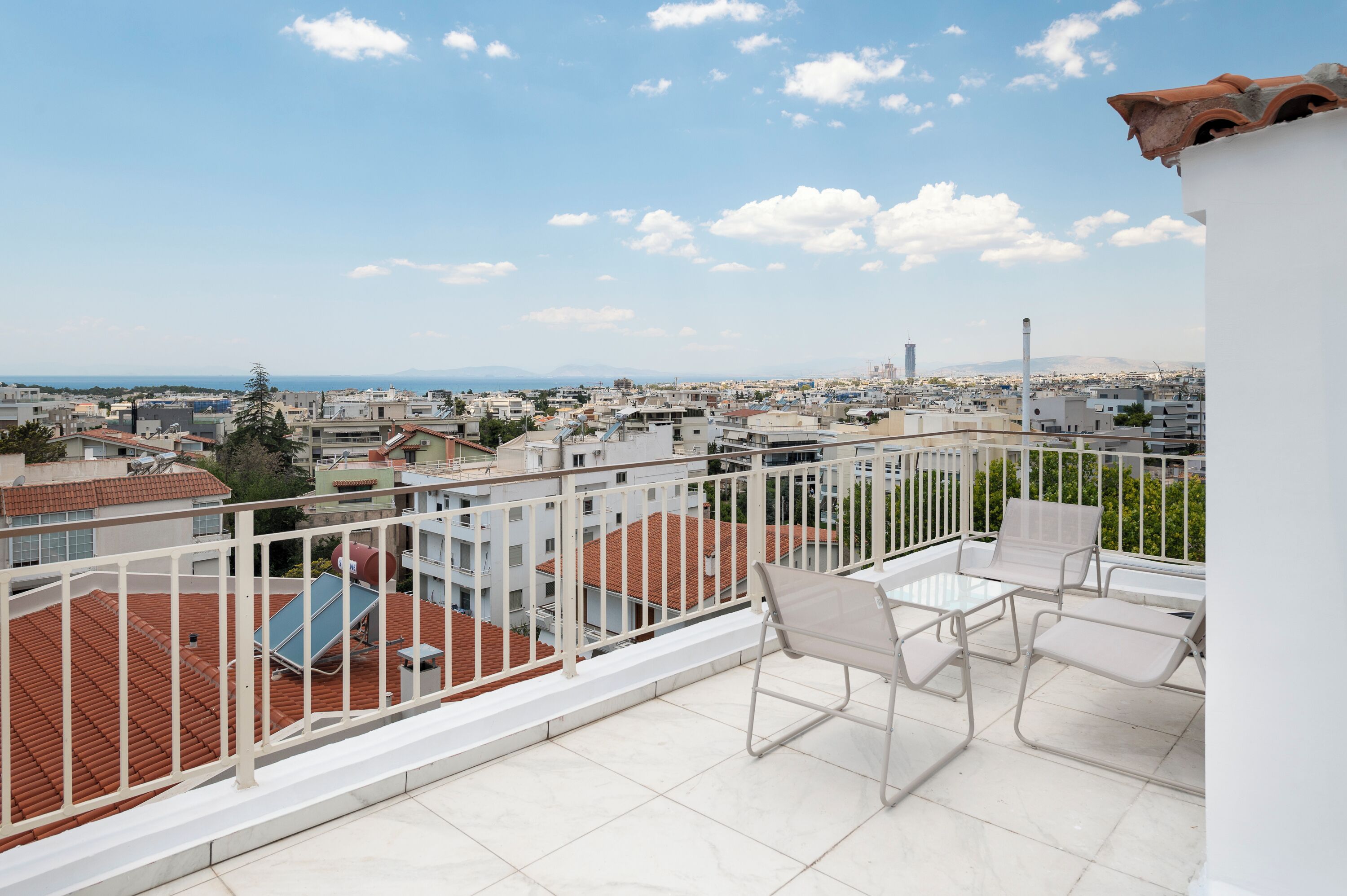 A balcony with white chairs and a table, overlooking a cityscape with buildings and a distant view of the sea.