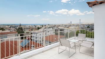 Un balcón con sillas y una mesa blancos, con vistas a un paisaje urbano con edificios y una vista distante del mar.