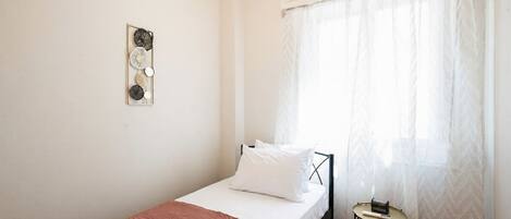 Una cama individual con colchón blanco y manta marrón, una mesita de noche con un libro y un objeto pequeño, y un adorno de pared.