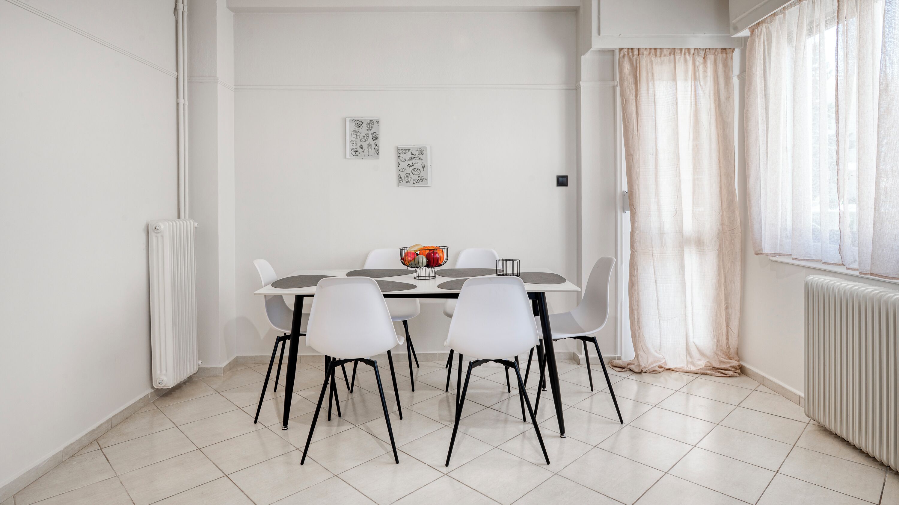 A dining area with a white table, four chairs, a bowl of fruit, and two wall decorations.