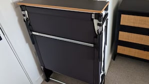 Ein kompakter, dunkel gefärbter, zweistufiger Stauraum mit Holzbeschichtung, platziert an einer weißen Wand in einem Raum mit Teppichboden.