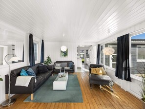 A modern living room with a black leather sofa, a glass coffee table, and a grey armchair. There is a large window on one side and a door leading to another room. - 4 Star Holiday Home in Lokken (Lokken)