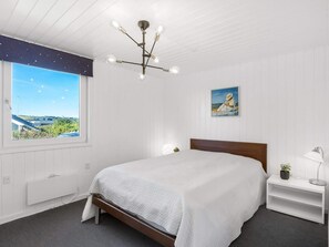Ein Schlafzimmer mit einem Bett, einem Nachttisch, einer Lampe, einem Gemälde und Blick auf eine Landschaft durch ein Fenster.
