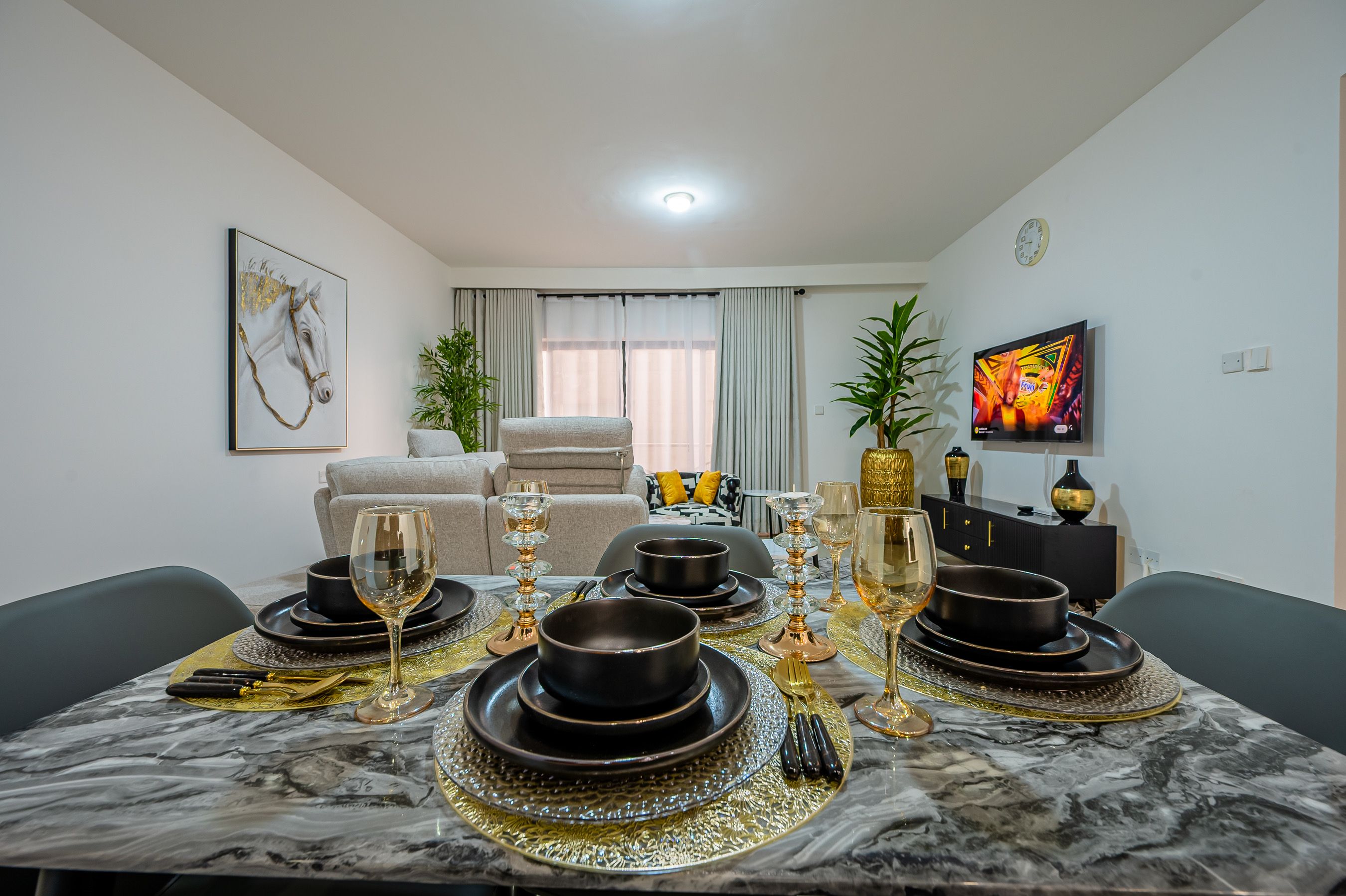 A dining area with a marble table set for four, a sofa, a TV, and a window with curtains.