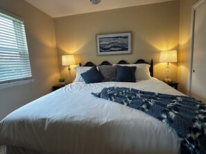 A neatly made bed with a patterned blanket, two bedside lamps, a framed picture on the wall, and a window with blinds. - Family-Friendly 4BR Getaway Close to Beaches, Fairhope & Fishing (Summerdale)