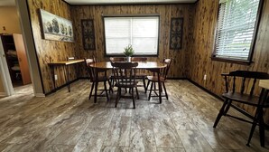 Een eethoek met een houten tafel en stoelen, een raam met jaloezieën, en een deur naar een andere kamer.