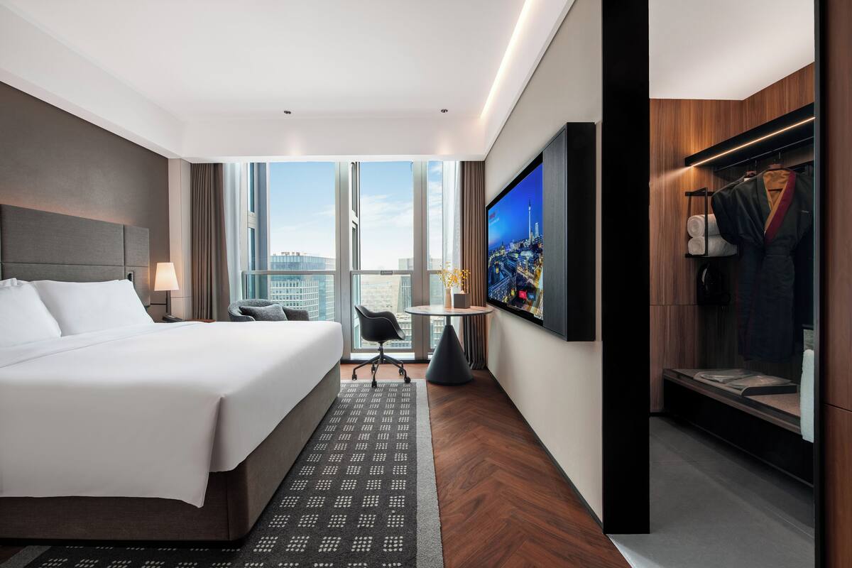 Ein modernes Hotelzimmer mit einem großen Bett, einem Fernseher, einer Sitzecke und Blick auf die Stadt.