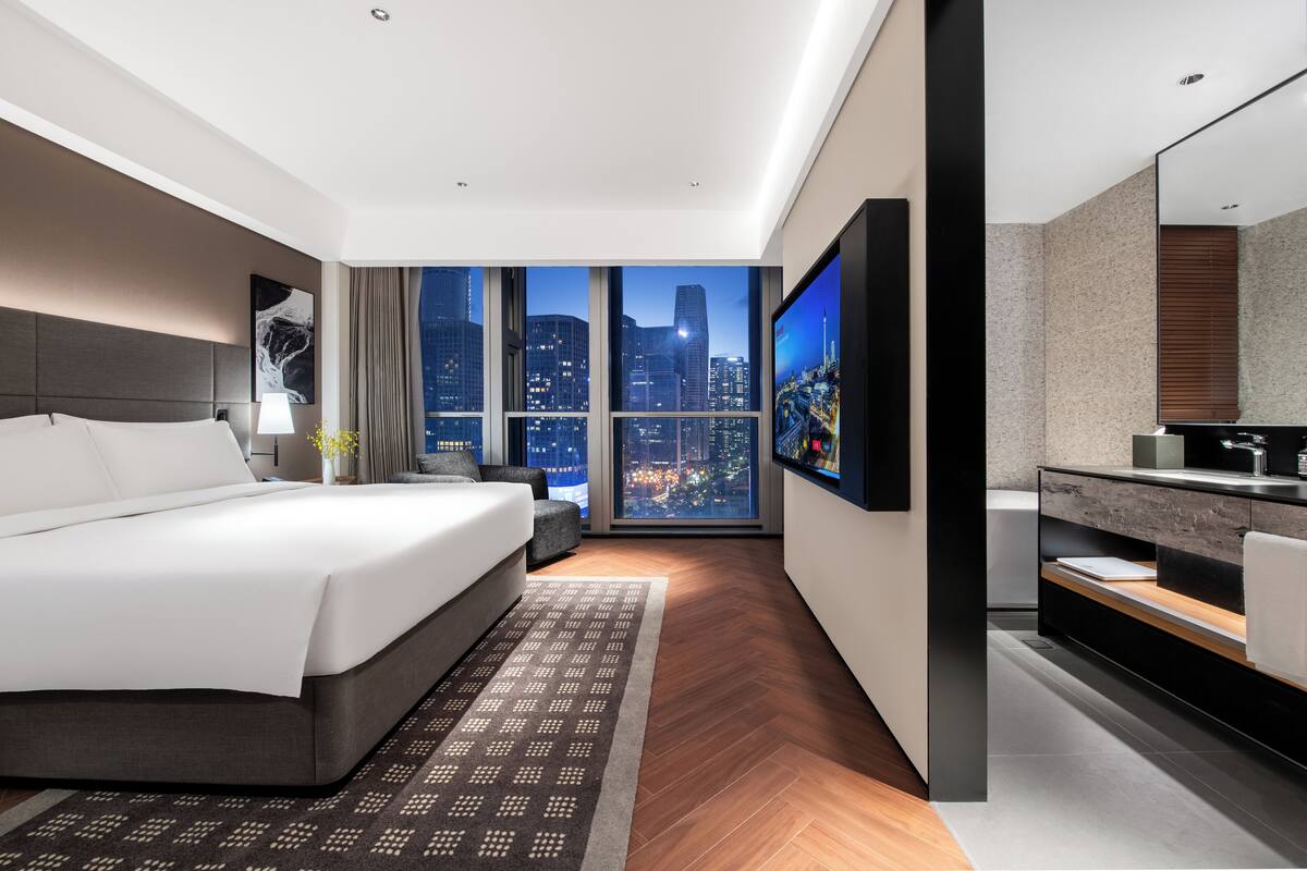 Ein modernes Hotelzimmer mit einem großen Bett, einem Fernseher, einem Badezimmer mit Spiegel und Blick auf die Stadt.