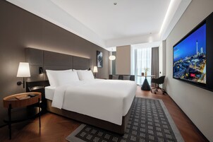 A modern hotel room with a large bed, a TV displaying a cityscape, a desk with a lamp, and a view of the city through large windows.