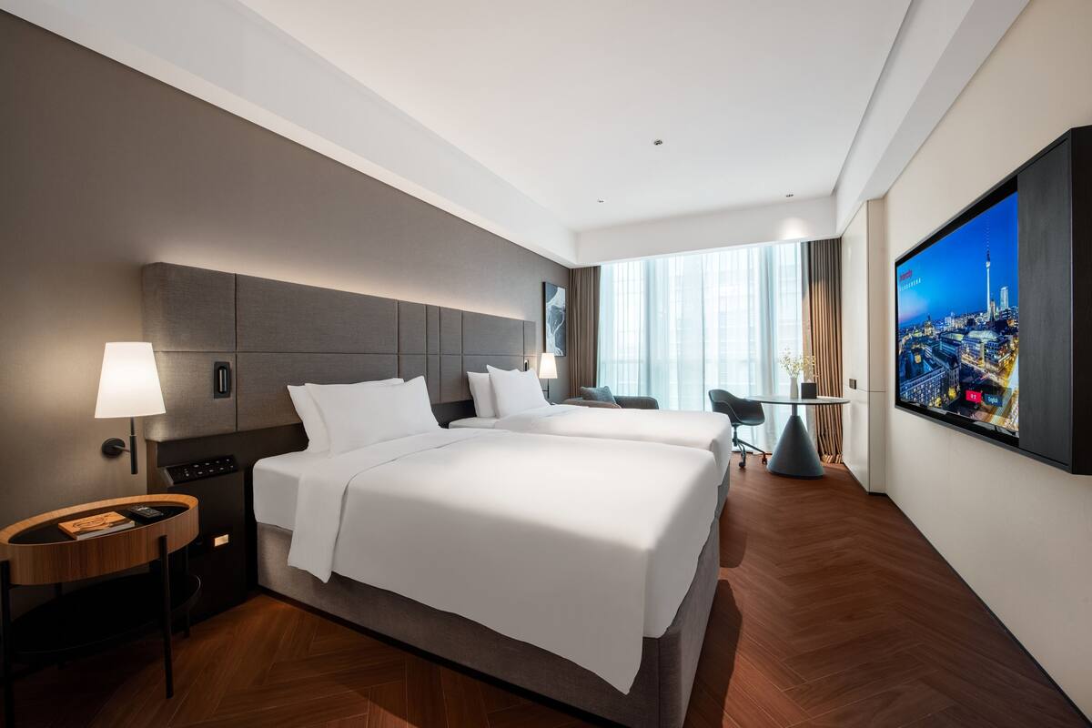 Ein modernes Hotelzimmer mit zwei Betten, einem Schreibtisch, einem Stuhl und einem großen Fernseher zur Stadtansicht.