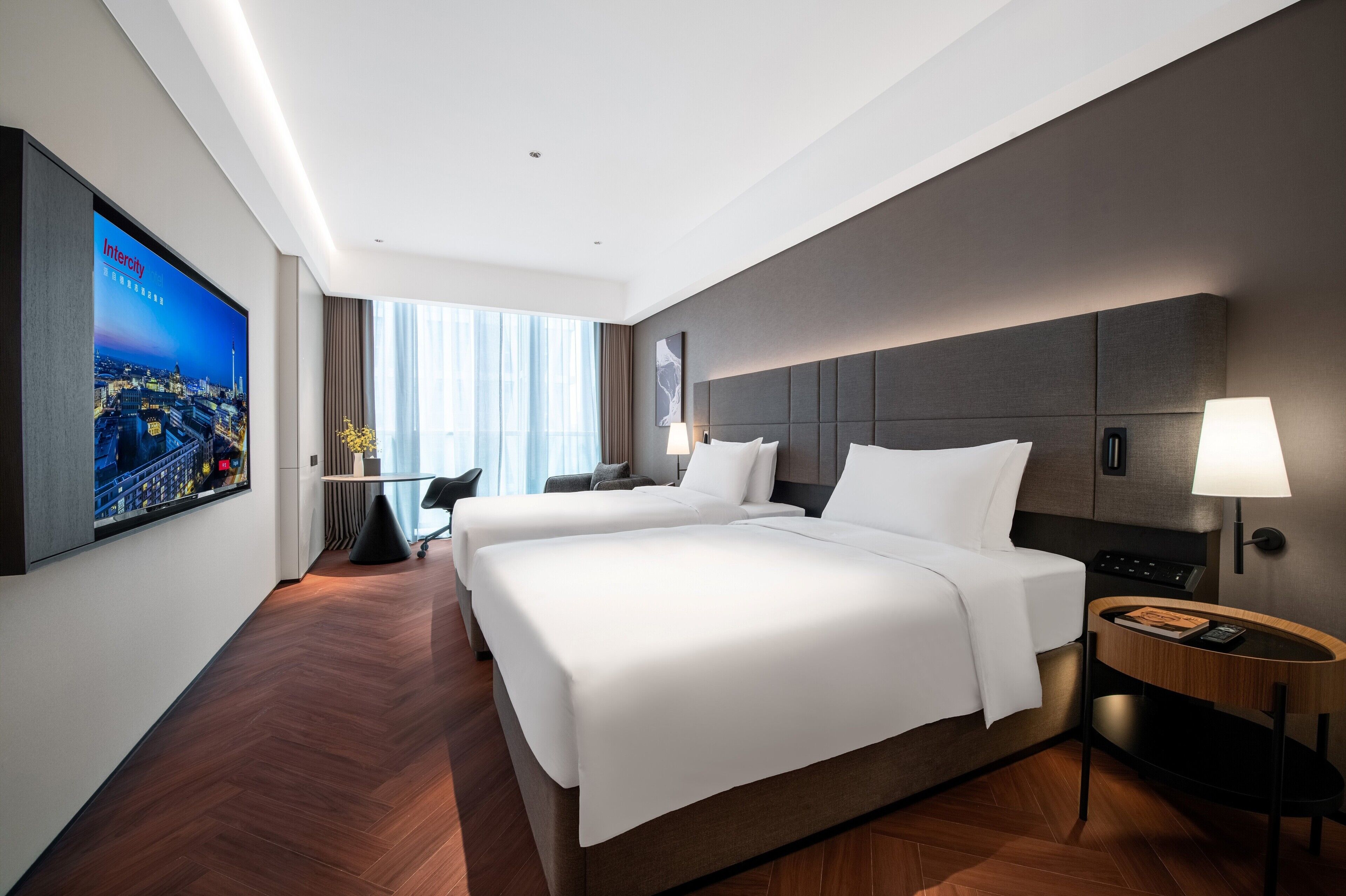 A hotel room with two beds, a TV on the wall, a desk, and a window with curtains.