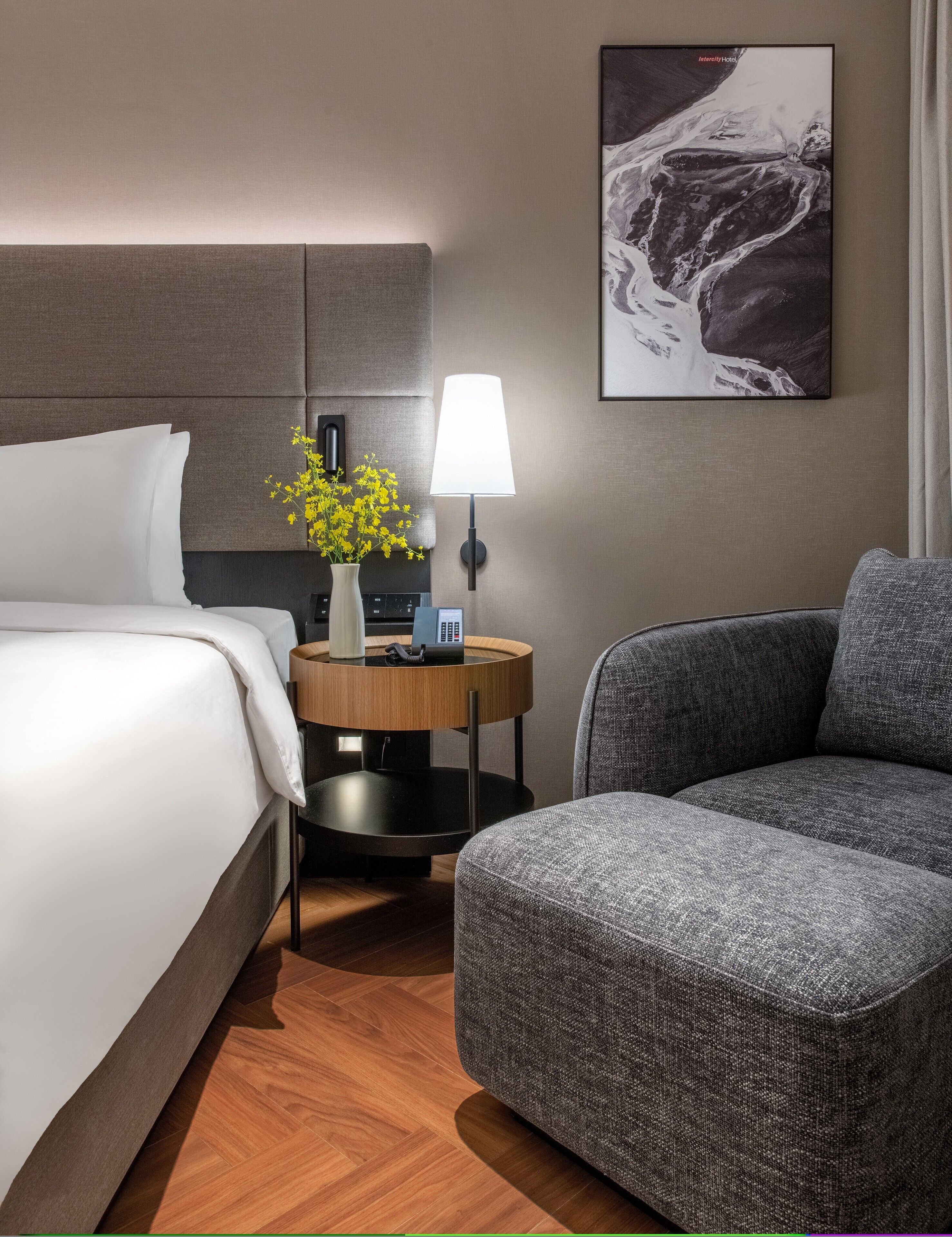 A hotel room with a bed, a chair, a bedside table with a lamp, a vase with flowers, a painting on the wall, and a telephone.