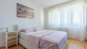 Un dormitorio con cama, mesita de noche, lámpara, un cuadro en la pared y una ventana con cortinas translúcidas.