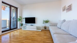 Una sala de estar con un televisor de pantalla plana, un sofá blanco, dos plantas en macetas y un ventanal corredizo que da a un balcón.