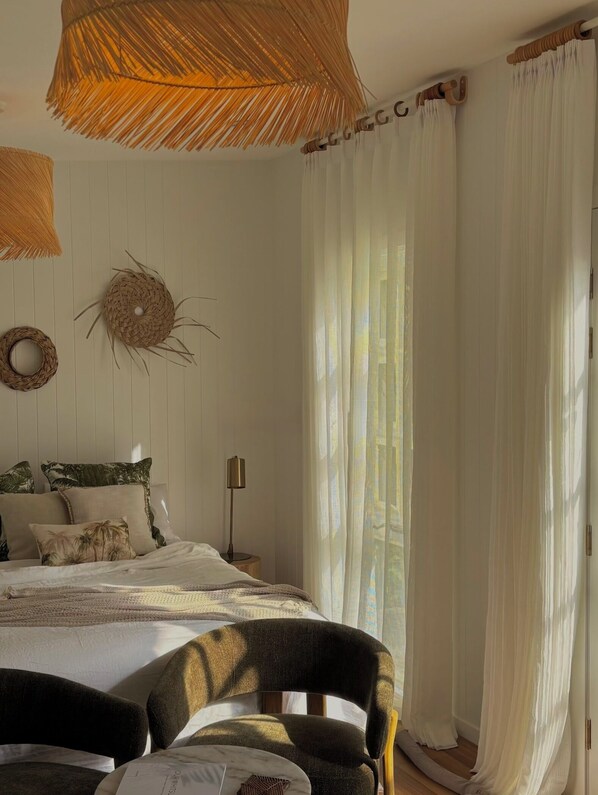 A bedroom with a bed, a nightstand, a lamp, a chair, and a large window with sheer curtains.