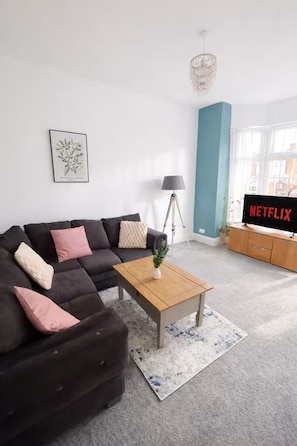 A living room with a sectional sofa, a coffee table, a TV, a floor lamp, and a window with curtains. - 30% off Comfortable 4-Bed Home 8 Beds Pets Welcome Sleeps 9 (Leeds)