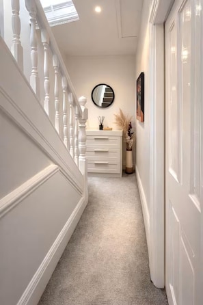 A narrow hallway with a white staircase, a round mirror on the wall, and a small white dresser with a vase of dried plants. - 30% off Comfortable 4-Bed Home 8 Beds Pets Welcome Sleeps 9 (Leeds)