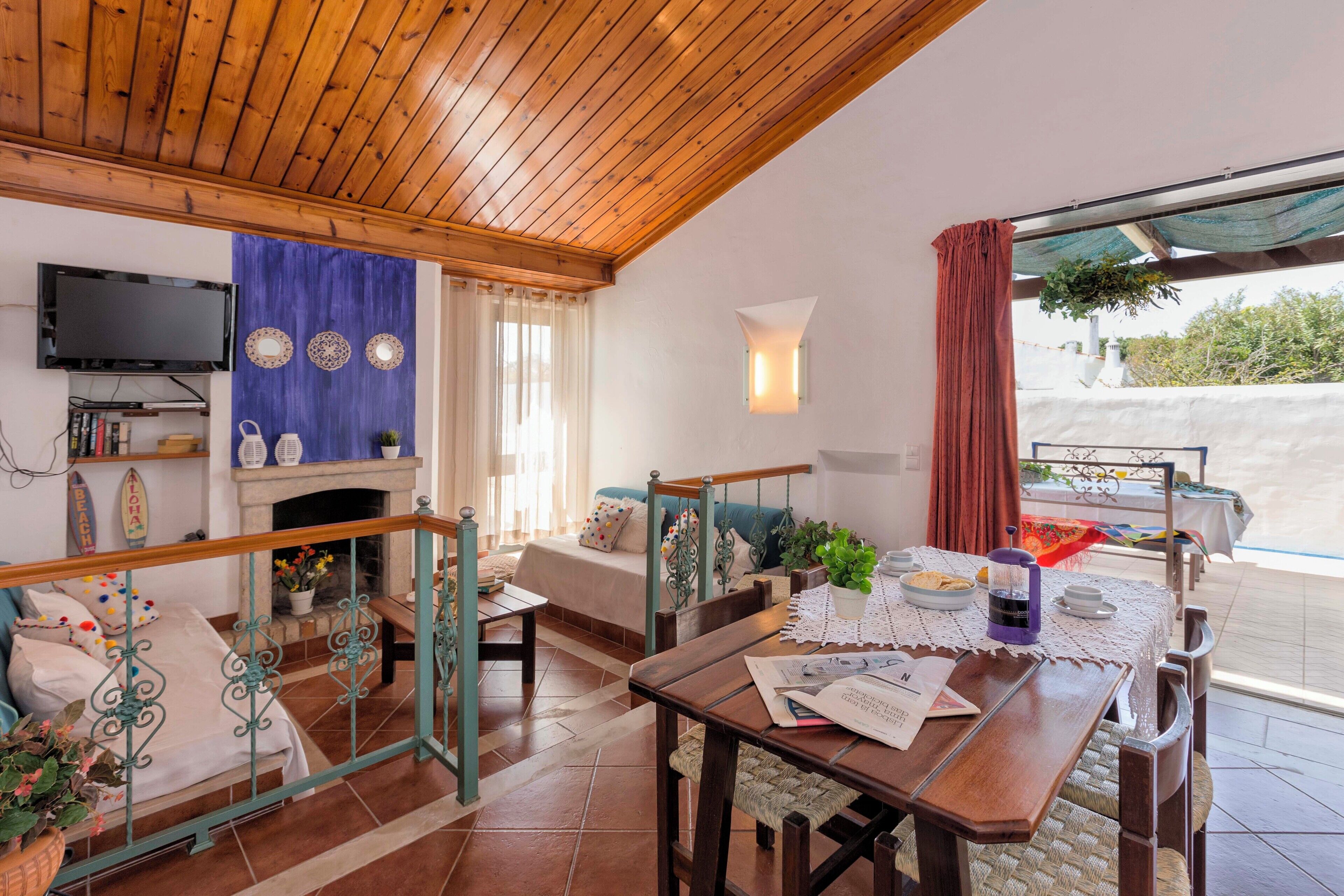 A cozy room with a wooden ceiling, a fireplace, a dining table with chairs, a bed with pillows, and a TV mounted on the wall.