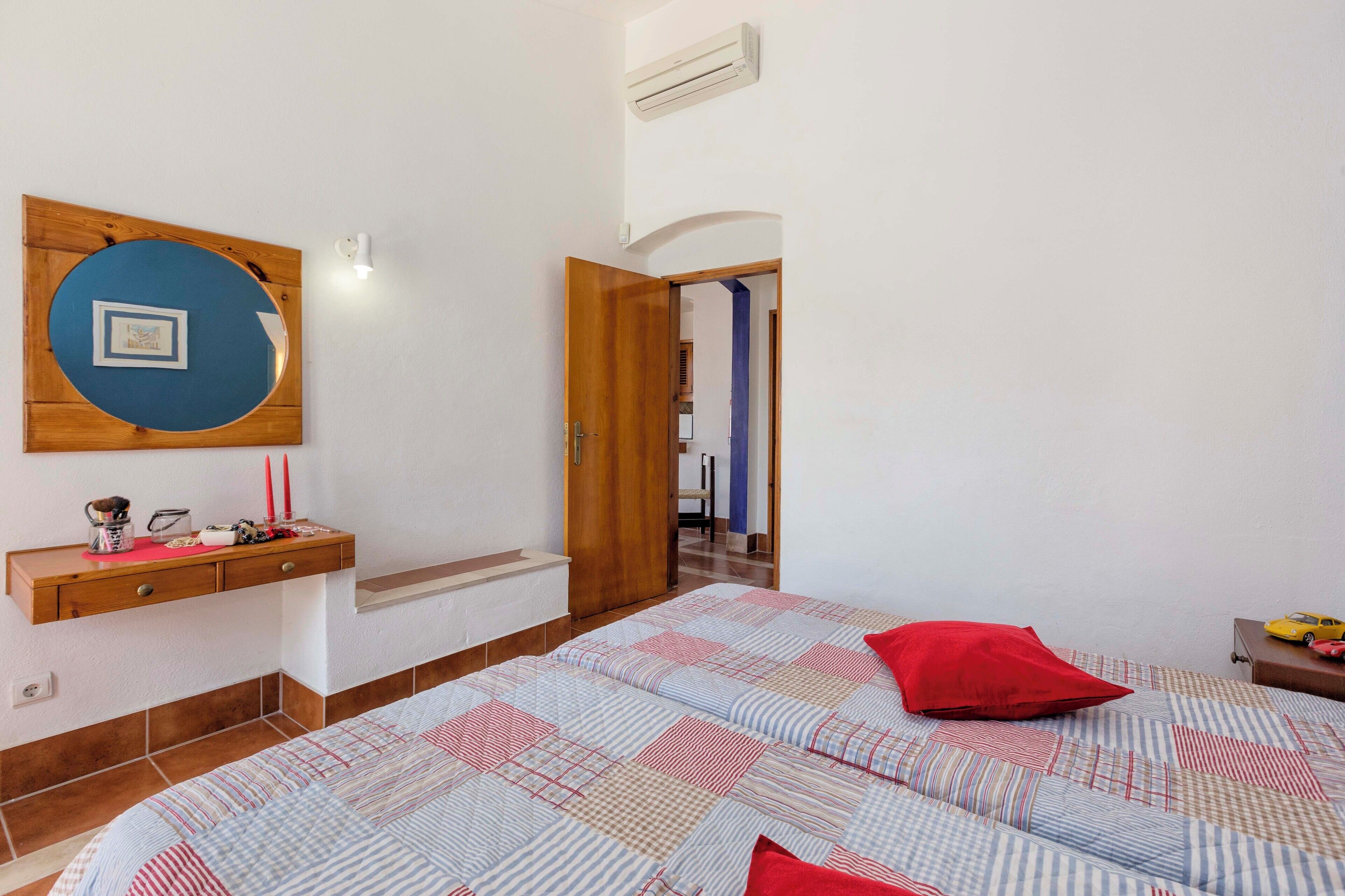 Un dormitorio con una cama, una mesita de noche de madera, un espejo adosado a la pared y una puerta que da a otra habitación.