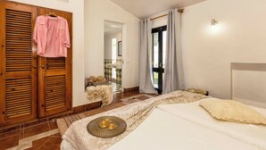 Ein Schlafzimmer mit einem Bett, einem Holzschrank, einem durchgehenden Türbogen sichtbaren Badezimmer und einem Fenster mit Vorhängen.