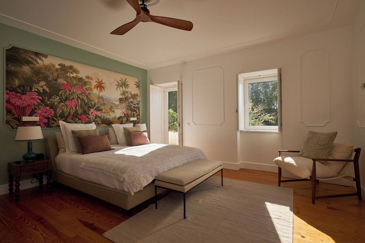 Une chambre à coucher avec un lit, une chaise, un ventilateur de plafond, une fenêtre et un tableau accroché au mur.
