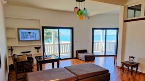 A spacious living area with a large sofa, a TV mounted on the wall, and a balcony with a view of the sea.