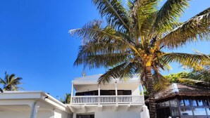 A two-story building with a balcony, surrounded by palm trees and a sandy area.