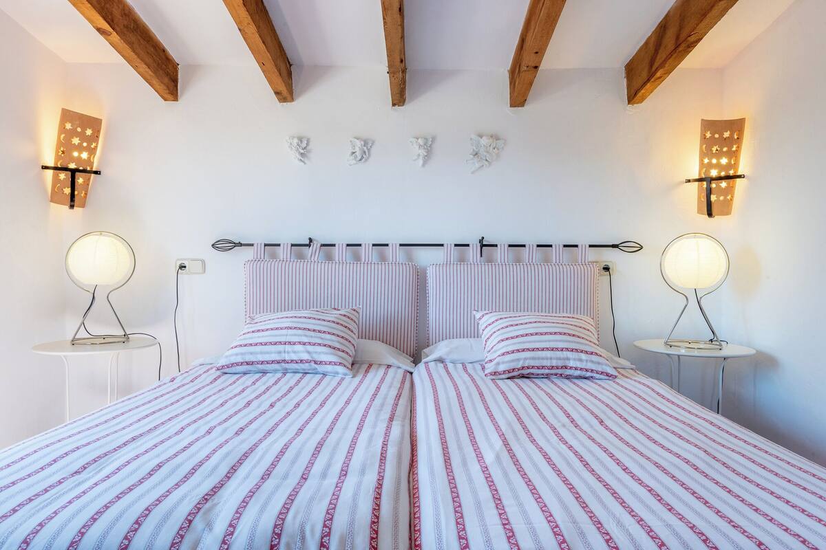 Un dormitorio con una cama, dos mesitas de noche, techo de madera y luces fijadas a la pared.
