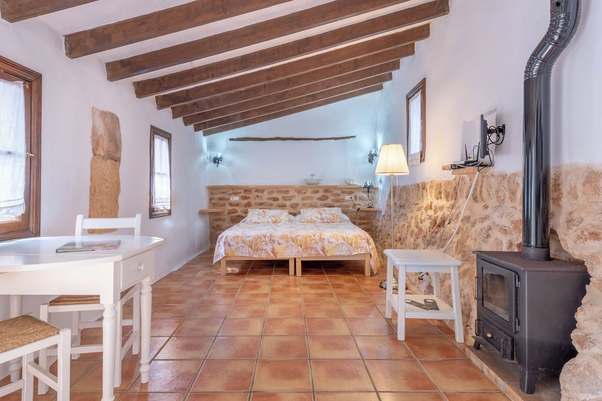 Un dormitorio rústico con pared de piedra, vigas de madera, cama, mesa, sillas, lámpara y estufa de leña.