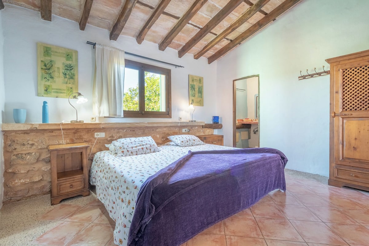 Un dormitorio con pared de piedra, vigas de madera, cama con ropa de cama floral, ventana con cortinas y un armario de madera.