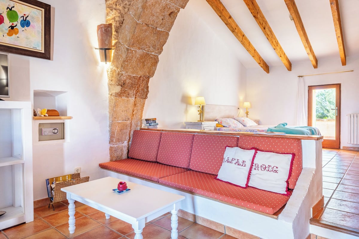 Una habitación acogedora con un arco de piedra, un sofá rojo con almohadas blancas, una mesa blanca y una cama con una colcha azul.