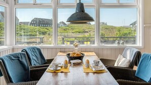 A dining area with wicker chairs, a wooden table, and a view of a coastal landscape through large windows. - 4 Star Holiday Home in Lokken (Lokken)
