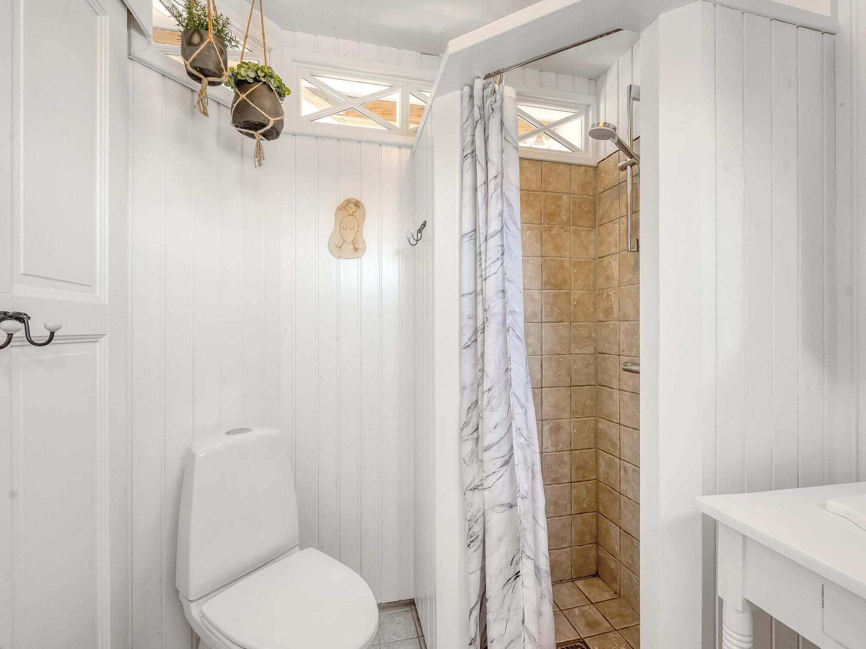 A bathroom with a white toilet, a shower with a marble curtain, and hanging plants.