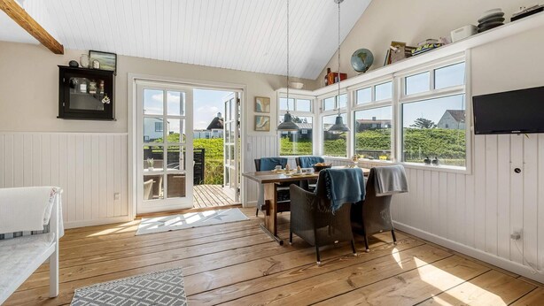 A room with a wooden floor, a dining table with chairs, a TV mounted on the wall, and a large window with a view of greenery and buildings. - 4 Star Holiday Home in Lokken (Lokken)