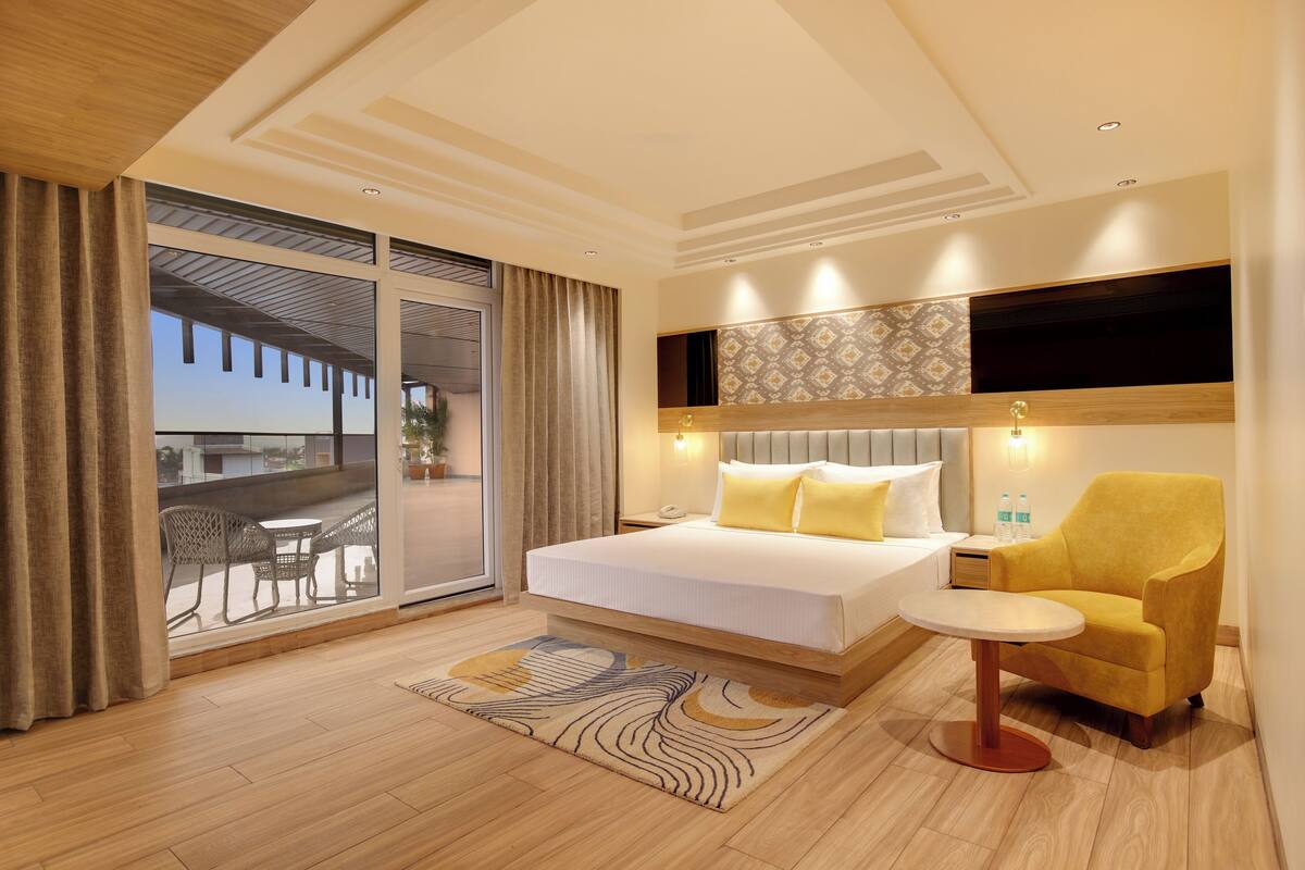 A hotel room with a bed, a chair, a small table, a balcony with outdoor furniture, and a patterned wall.