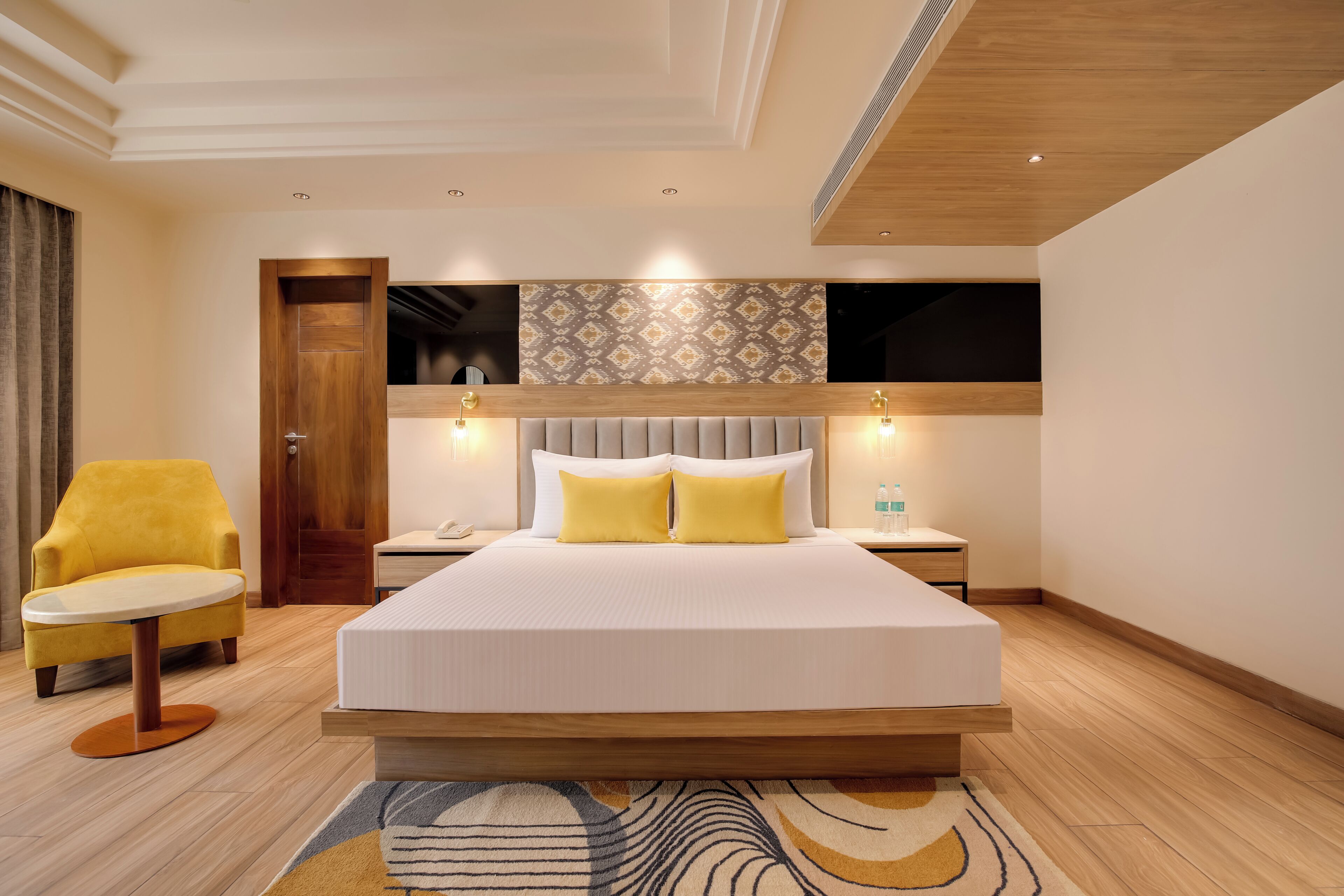 Une chambre d’hôtel moderne avec un grand lit, un fauteuil jaune, une table de chevet et un téléviseur fixé au mur.